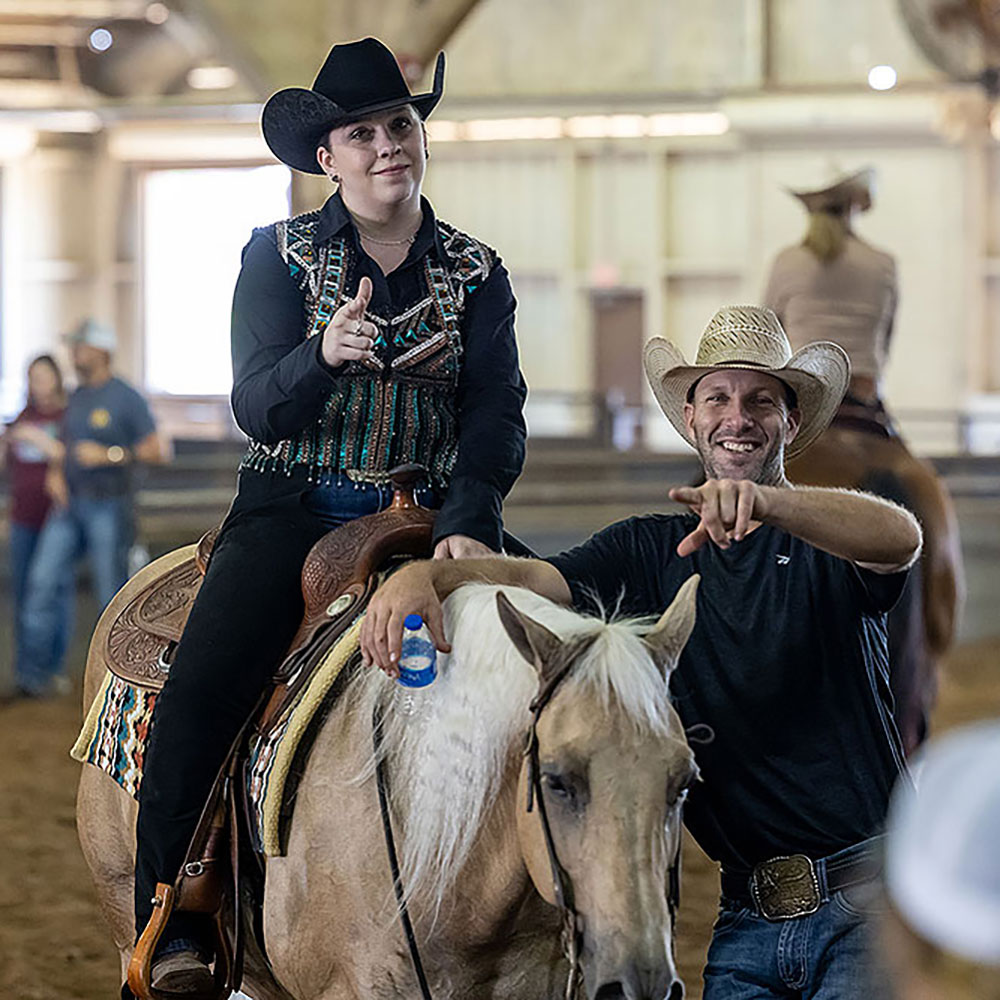 Woman in western outfit on a horse and man in cowboy hat smiling and pointing at camera.