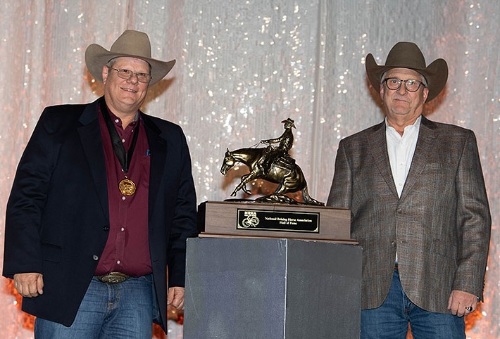 Two men with cowboy hats and medals, standing beside a cowboy riding trophy, on stage with NRHA Hall of Fame backdrop.